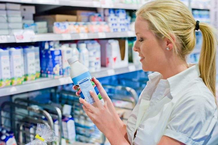 Mujer rubia eyendo etiquetado de envase de leche de soja del lineal del supermercado